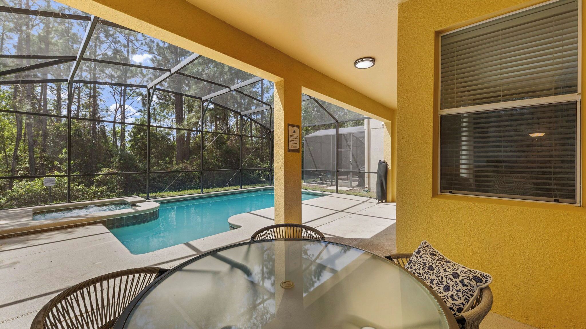 A glass-enclosed outdoor pool area with a hot tub, surrounded by a yellow wall and a view of a forest.