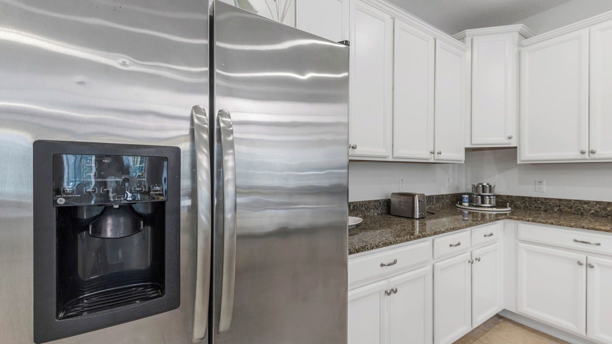 Un refrigerador de acero inoxidable con dispensador de agua, armarios blancos, encimera de granito y un tostador.