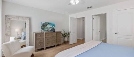 A bedroom with a bed, a dresser, a chair, a ceiling fan, and a painting on the wall.