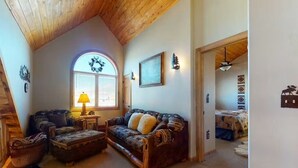 A cozy living room with a wooden ceiling, a large window, a sofa, a coffee table, and a lamp. - 4BR Riverfront | Hot Tub | Fireplace (South Fork)