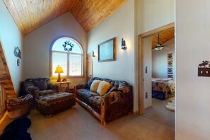 A cozy living room with a wooden ceiling, a large window, a sofa, a coffee table, and a lamp. - 4BR Riverfront | Hot Tub | Fireplace (South Fork)