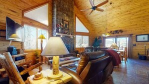 A cozy cabin interior with a stone fireplace, wooden walls, a leather sofa, and a ceiling fan. - 4BR Riverfront | Hot Tub | Fireplace (South Fork)