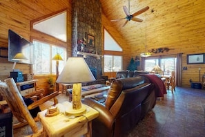 A cozy cabin interior with a stone fireplace, wooden walls, a leather sofa, and a ceiling fan. - 4BR Riverfront | Hot Tub | Fireplace (South Fork)