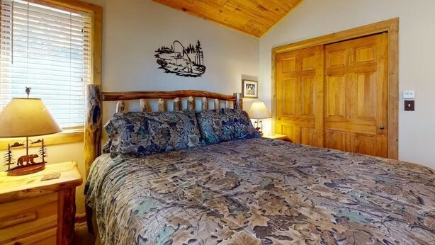 A bedroom with a wooden ceiling, a bed with patterned bedding, a nightstand with a lamp, and a wooden door. - 4BR Riverfront | Hot Tub | Fireplace (South Fork)
