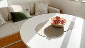 Une pièce avec une table ronde, un saladier rempli de fruits, un canapé avec des coussins et une fenêtre dont le volet est ouvert.
