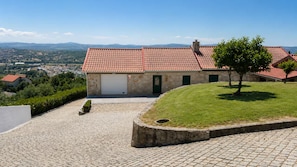 Una casa in pietra a due piani con tetto in tegole rosse, un garage e un vialetto lastricato che conduce a una zona verdeggiante con un albero.