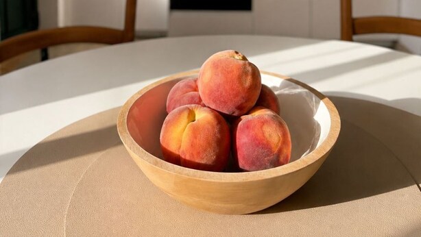 Une table à manger avec un saladier de fruits au premier plan, une cuisine en arrière-plan avec un four et des placards, et deux chaises en bois.