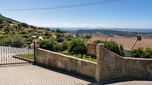 Un muro di pietra con un cancello in metallo, un cartello che riporta 'CASA Entre CAMPO' e vista su un borgo con case e colline all'orizzonte.