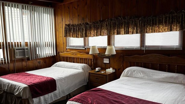 Een hotelkamer met twee bedden, houten lambrisering, een raam met zonwering en een nachtkastje met een lamp.