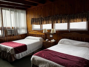 A hotel room with two beds, wooden paneling, a window with blinds, and a nightstand with a lamp.