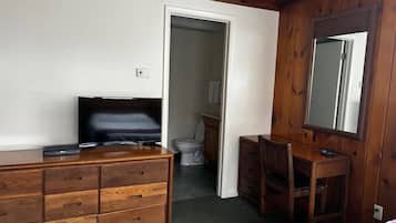 A bedroom with a wooden dresser, a television, a desk, and a mirror. There is a door leading to a bathroom.