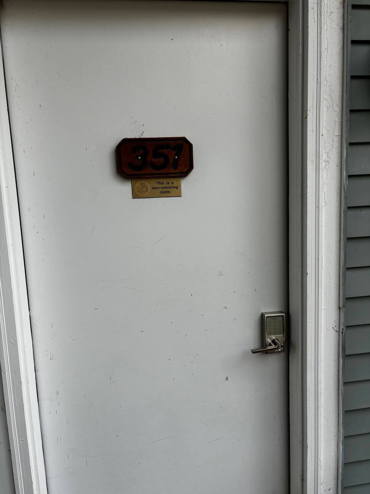 Une porte blanche ornée d’une plaque en bois portant le numéro 357 et, en dessous, d’un panneau indiquant qu’il est interdit de fumer.