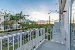Un balcon doté d’une balustrade blanche, donnant sur une rue où sont garées des voitures et où se trouvent des immeubles.