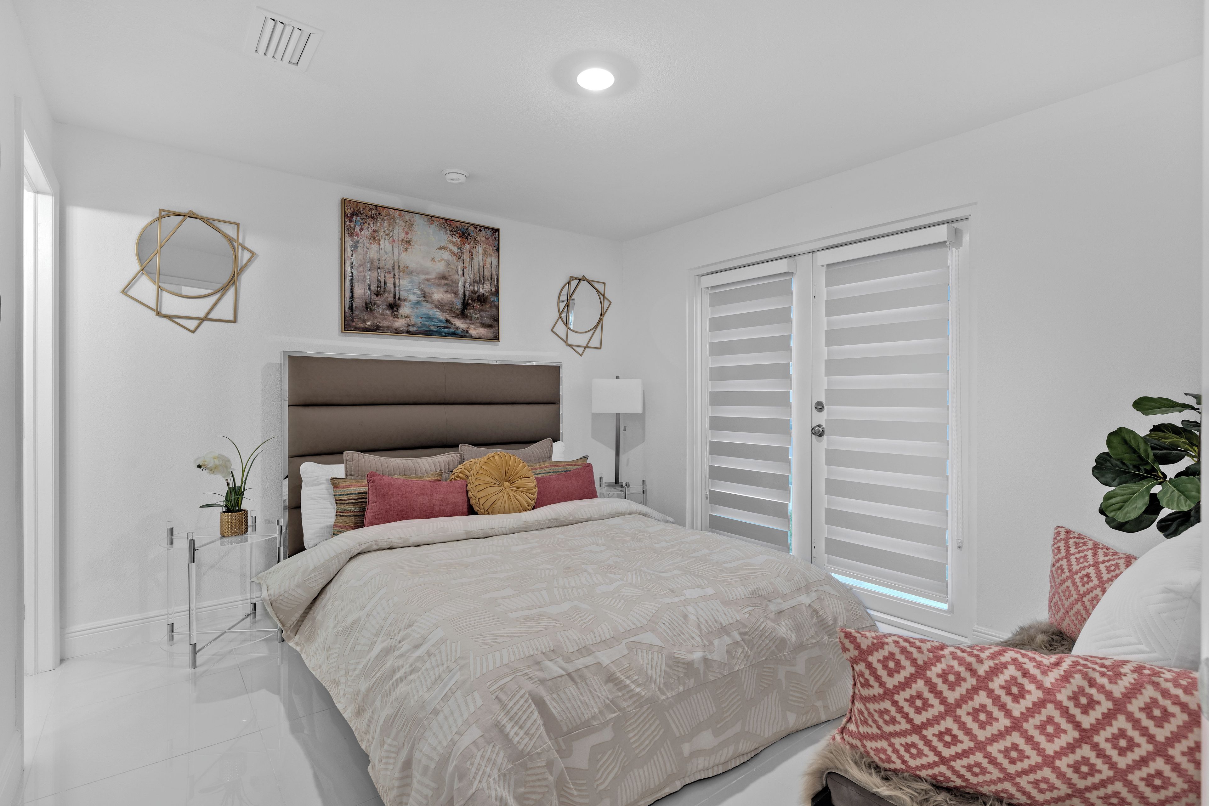 A bedroom with a bed, a nightstand, a bedside table, a window with blinds, a painting on the wall, and a plant.