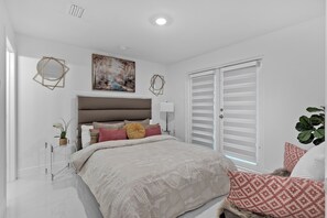 Un dormitorio con cama, mesita de noche, ventana con persianas, un cuadro en la pared y una planta.
