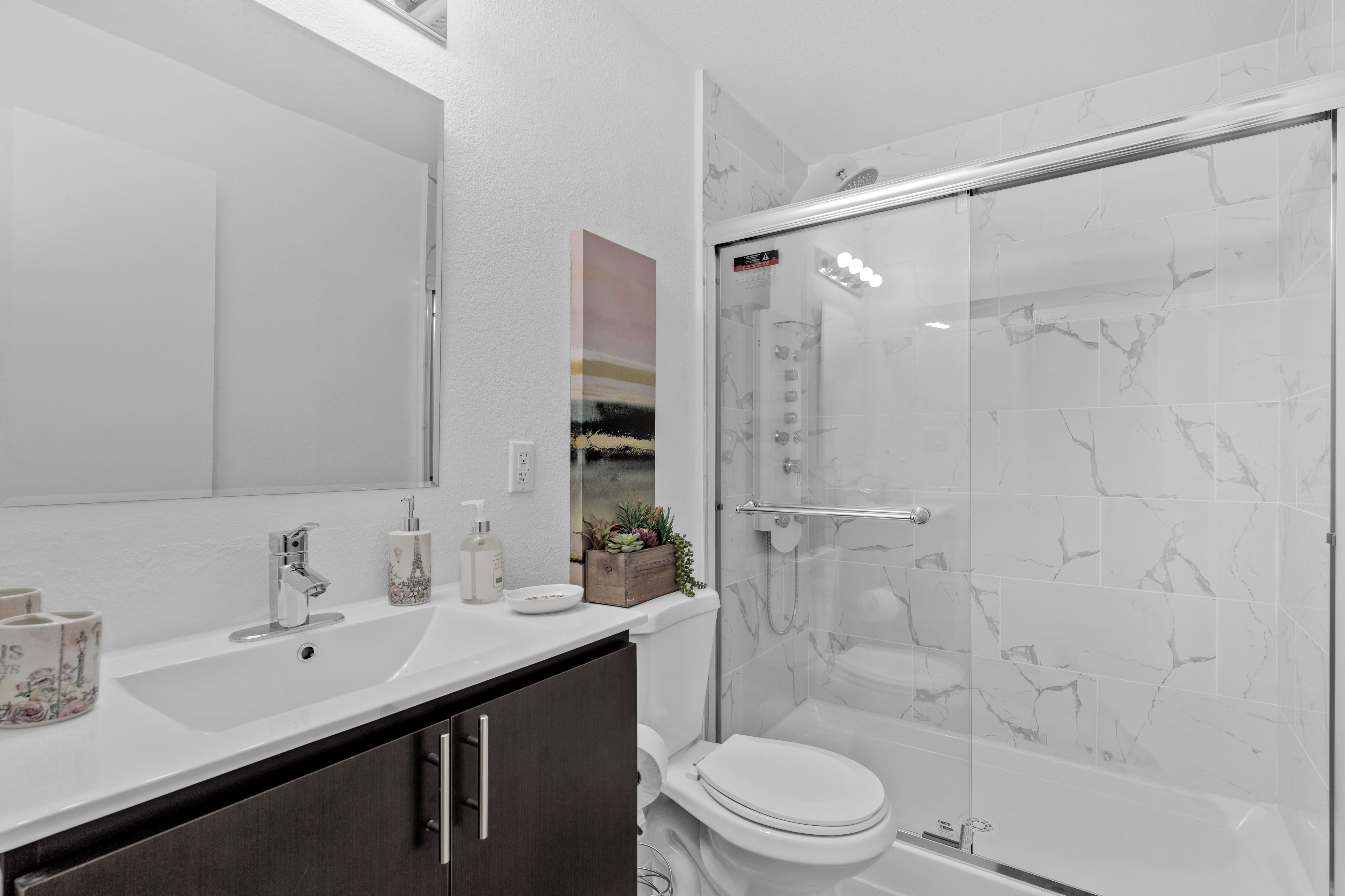 A modern bathroom with a white sink, dark wooden cabinets, a glass shower enclosure, and a marble-tiled shower wall.