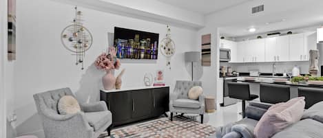 A modern living room with a grey sofa, a grey armchair, and a patterned rug. There is a flat-screen TV mounted on the wall, a decorative wall piece, and a small table with a vase and books.