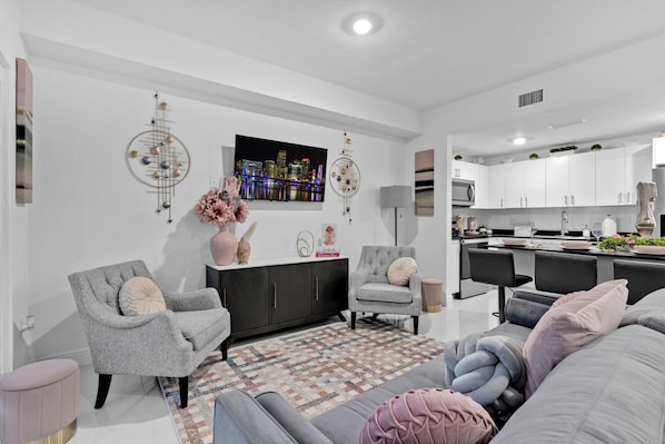 Un salon moderne avec un canapé gris, un fauteuil gris et un tapis à motifs. Un téléviseur à écran plat est fixé au mur, ainsi qu’un élément décoratif mural et une petite table sur laquelle se trouvent un vase et des livres.