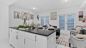 A modern kitchen with a central island, white cabinetry, and a dining area with a grey sofa and blue shutters. - Peaceful Dreamscape a spacious family friendly minutes to the Keys & Miami (Princeton)