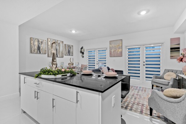 A modern kitchen with a central island, white cabinetry, and a dining area with a grey sofa and blue shutters.