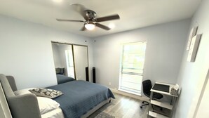 Ein Schlafzimmer mit Bett, Schreibtisch, Stuhl, Deckenventilator, einem Fenster mit Jalousien und einer Tür.