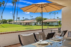 A dining table with chairs and a large umbrella, overlooking a golf course with palm trees and buildings in the background. - Gated Golf Course Condo at Rancho Las Palmas – Fairway & Mountain Views, Resort Pools & Tennis (Rancho Mirage)