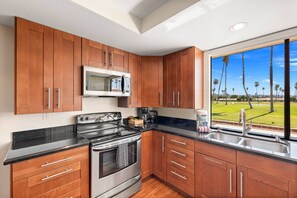 A kitchen with wooden cabinets, a stainless steel stove, a microwave, and a window with a view of palm trees. - Gated Golf Course Condo at Rancho Las Palmas – Fairway & Mountain Views, Resort Pools & Tennis (Rancho Mirage)