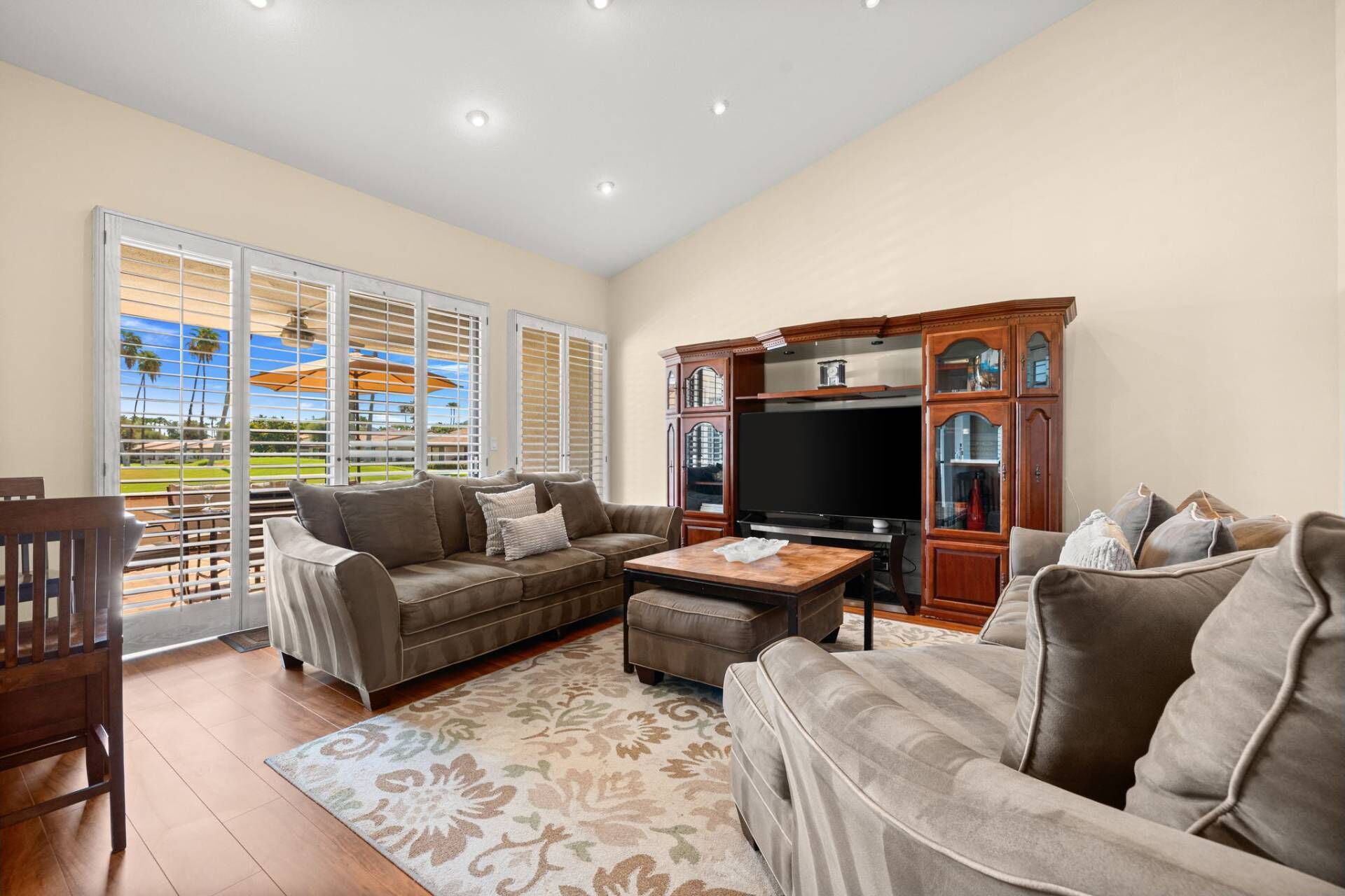 A spacious living room with a large flat-screen TV, a brown leather sofa, a matching ottoman, and a wooden cabinet with glass doors.