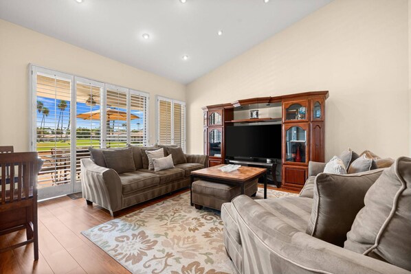 A spacious living room with a large flat-screen TV, a brown leather sofa, a matching ottoman, and a wooden cabinet with glass doors.