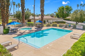 A rectangular swimming pool with a diving board, surrounded by palm trees and a fenced area with lounge chairs and umbrellas.