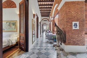 A hotel corridor with a checkered floor, a bed with white linens, a wooden door, and a brick wall with a hand symbol. - Grand Historic Mansion in Old SJ | Patio + Rooftop (San Juan)
