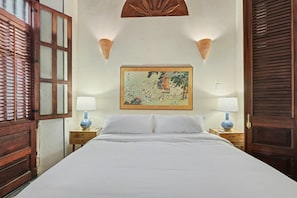 A hotel room with a large bed, two bedside tables with lamps, a wall-mounted painting, and wooden shutters. - Grand Historic Mansion in Old SJ | Patio + Rooftop (San Juan)