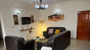 Ein Wohnzimmer mit einem schwarzen Ledersofa, einem weißen Sessel und einem Couchtisch. An der Wand ist ein Fernseher angebracht, außerdem steht eine Lampe und ein Gemälde von Pferden dort.