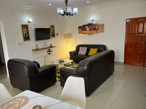 Un salón con un sofá de cuero negro, un sillón blanco y una mesa de centro. Hay un televisor montado en la pared, una lámpara y un cuadro de caballos.