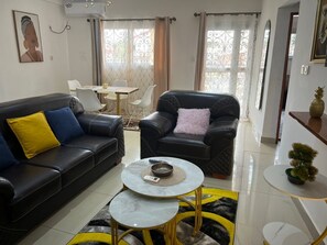 Ein Wohnzimmer mit einer schwarzen Couch, einem schwarzen Sessel, einem runden Couchtisch und einem Essbereich mit einem weißen Tisch und Stühlen.