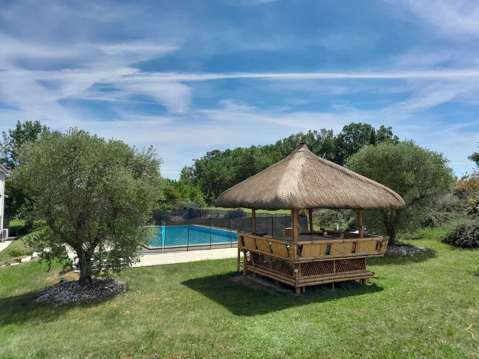 Un espace piscine agrémenté d’un gazebo à toit de chaume et d’un arbre.