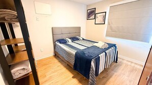 Un dormitorio con cama, piso de madera, cabecero, ventana con persianas y un interruptor de luz fijado a la pared.