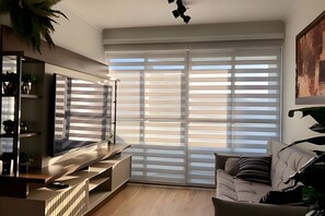 Una sala de estar moderna con un televisor de pantalla plana, un sofá y un ventanal con persianas.