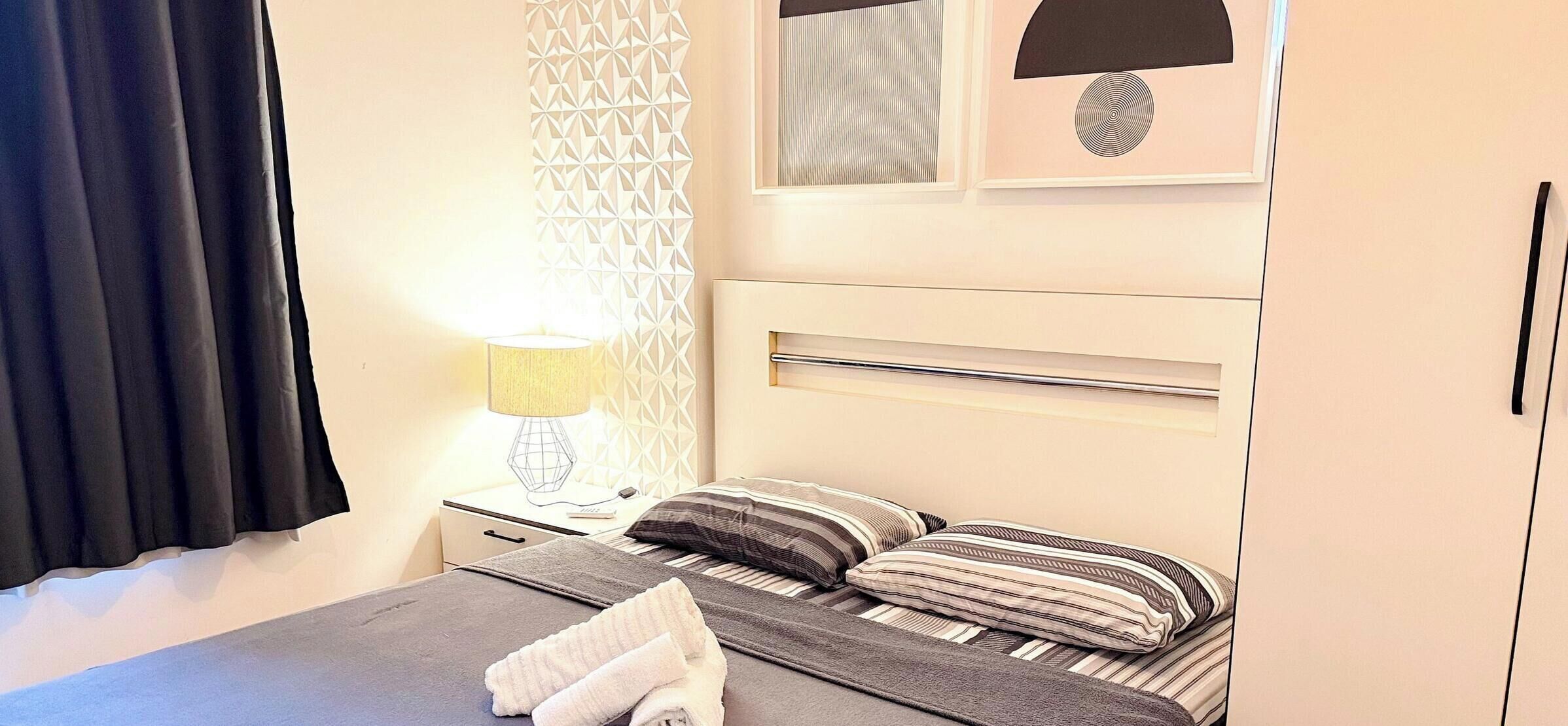 A neatly made bed with pillows and a folded towel, a bedside lamp, and a wall with a geometric pattern.