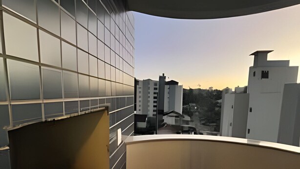 A balcony with a view of buildings, a tiled wall, and a metal grill. - ASA Apartamento Cartagena Oktoberfest (Blumenau)