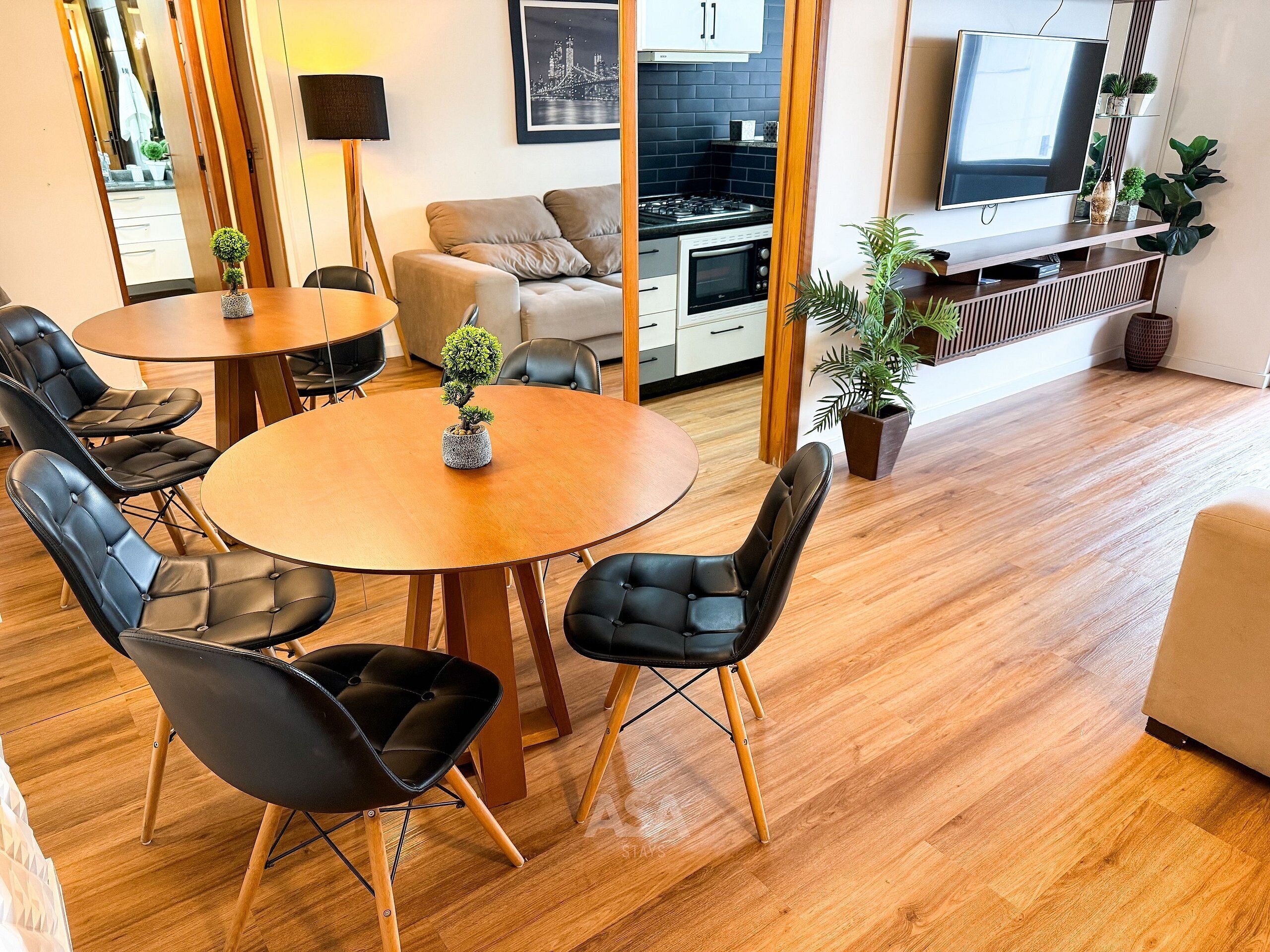 Ett modernt vardagsrum med ett runt träbord, svarta läderstolar, en soffa, en köksdel med en spis och ugn samt en tv på en hylla.