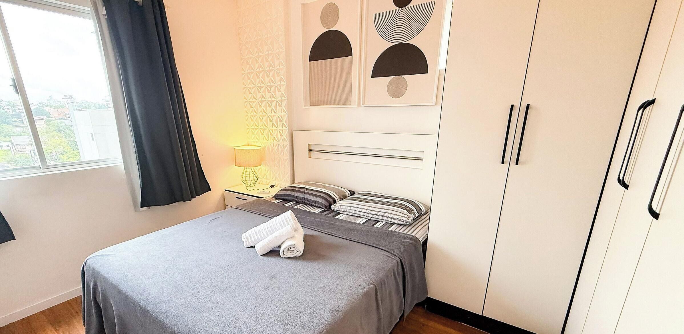 Um quarto com uma cama, mesa de cabeceira, abajur, guarda-roupa e janela com cortinas.