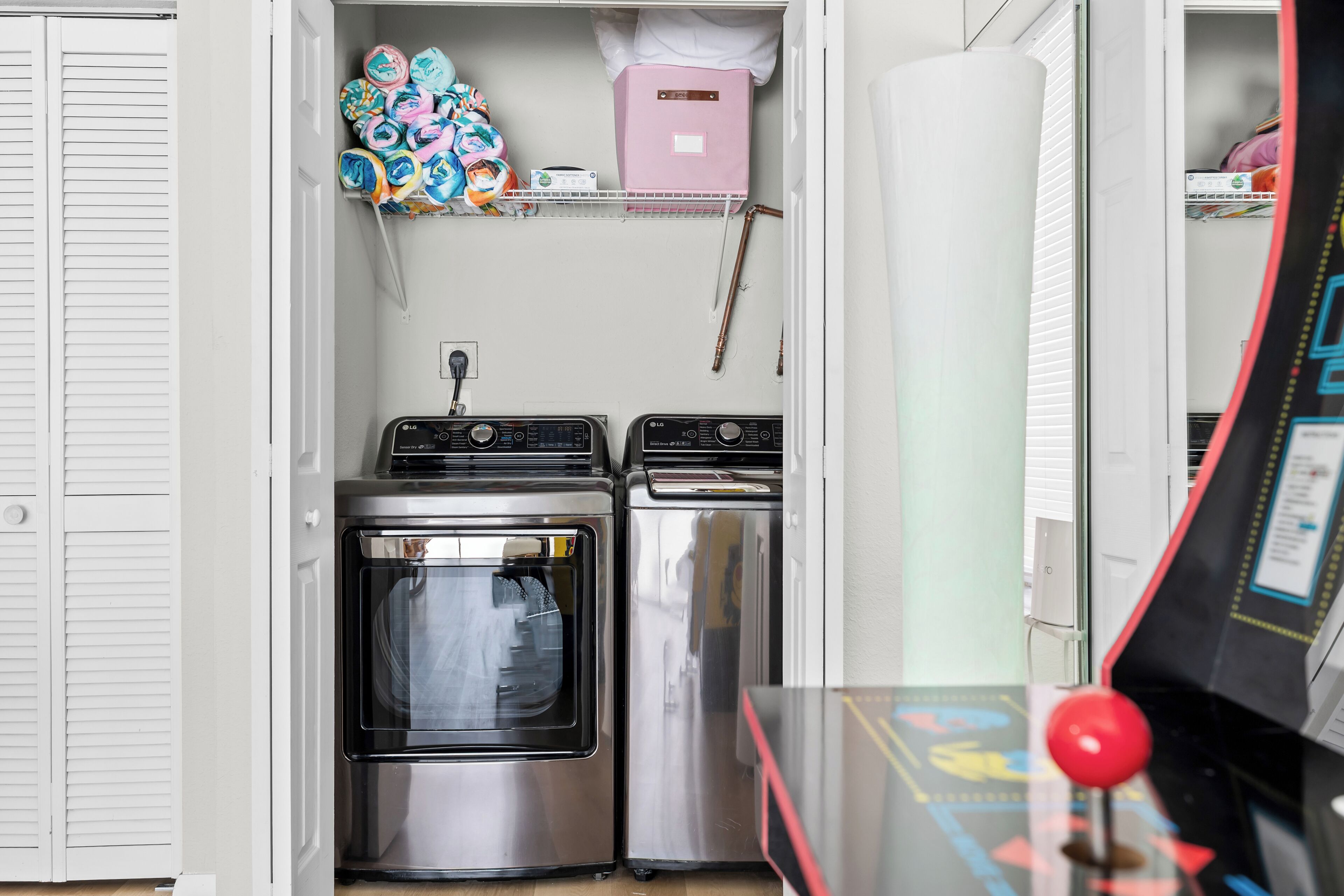A compact laundry room with two washing machines and a dryer, a shelf with folded towels, and a pink storage box.