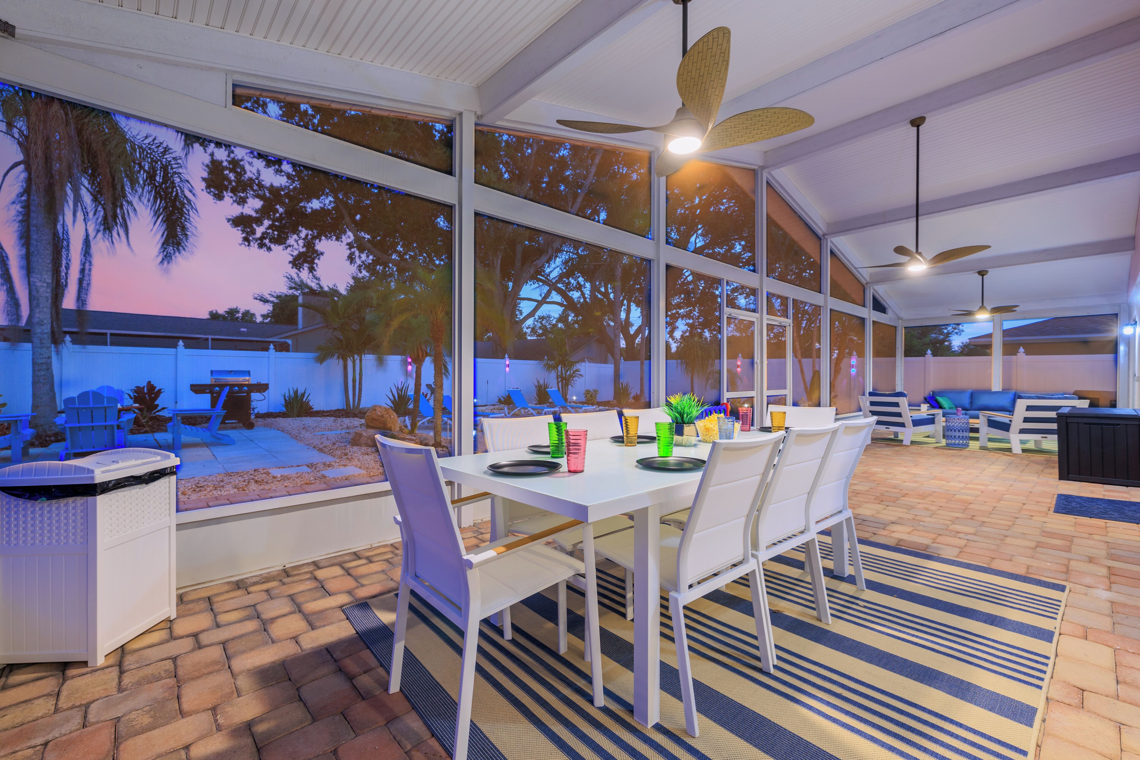 An outdoor dining area with a table set for two, surrounded by white chairs, and a view of the sky through large windows.