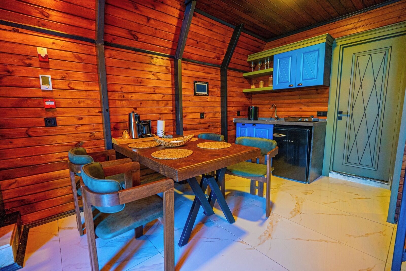 A wooden interior with a dining table, chairs, a kitchen area with blue cabinets, and a door with a geometric pattern.