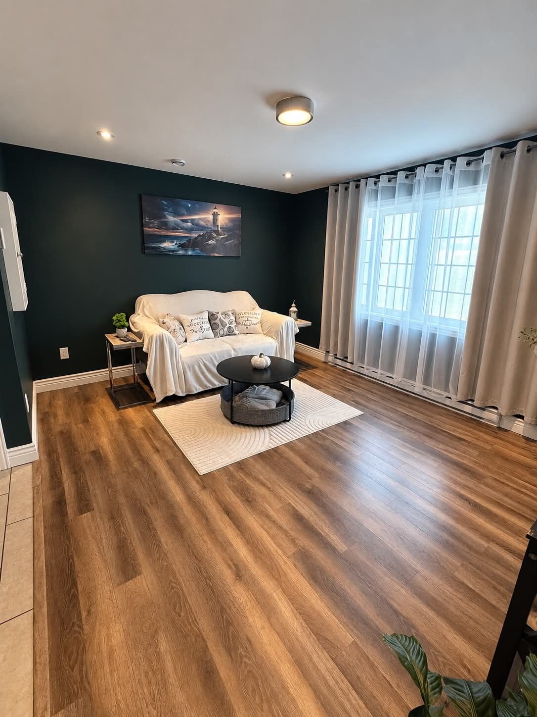Ein Wohnzimmer mit dunkler Wand, einem weißen Sofa, einem Couchtisch und einem Fenster mit Vorhängen.