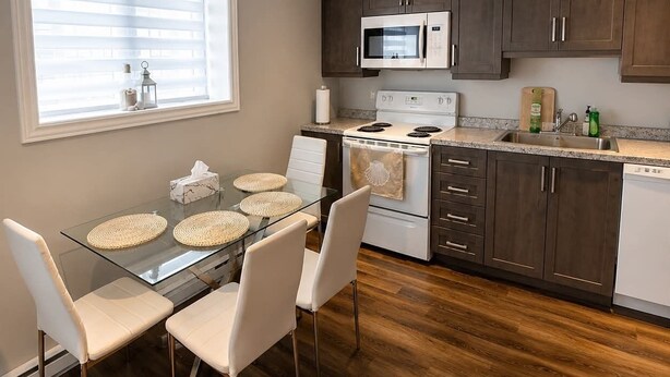 A modern kitchen with a glass dining table, white chairs, wooden flooring, and dark cabinets.