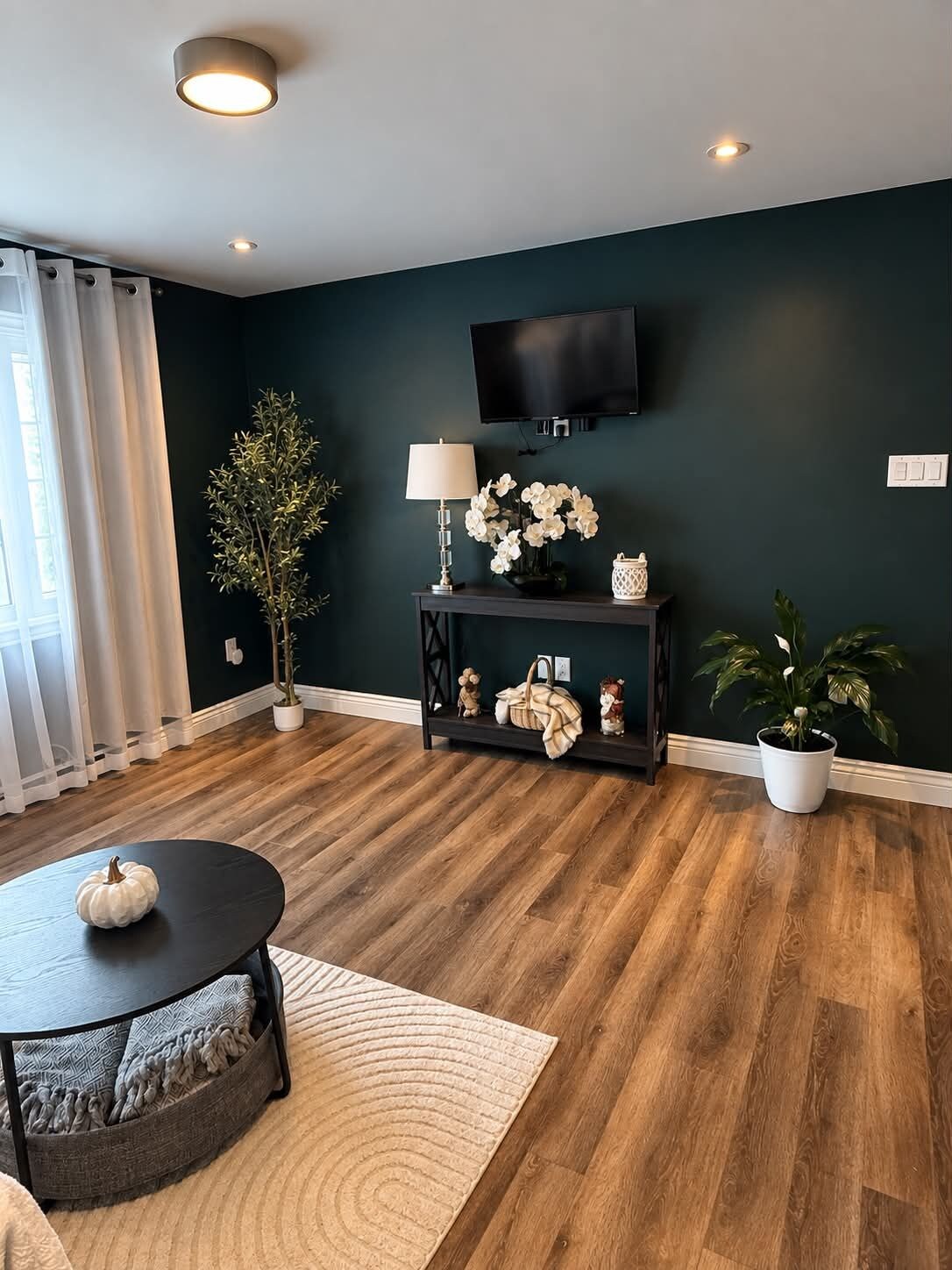 A modern living room with a dark wall, wooden flooring, a round coffee table, a TV mounted on the wall, and a potted plant.