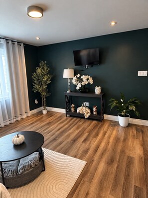 A modern living room with a dark wall, wooden flooring, a round coffee table, a TV mounted on the wall, and a potted plant. - Cozy 1BR Suite in Kenmount Terrace. (St. John's)