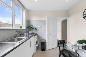 A modern kitchen with a built-in washing machine, a sink, and a dining area with a glass table. - Family & Contractor Stay | Sleep 7 |NR Heathrow (Langley)